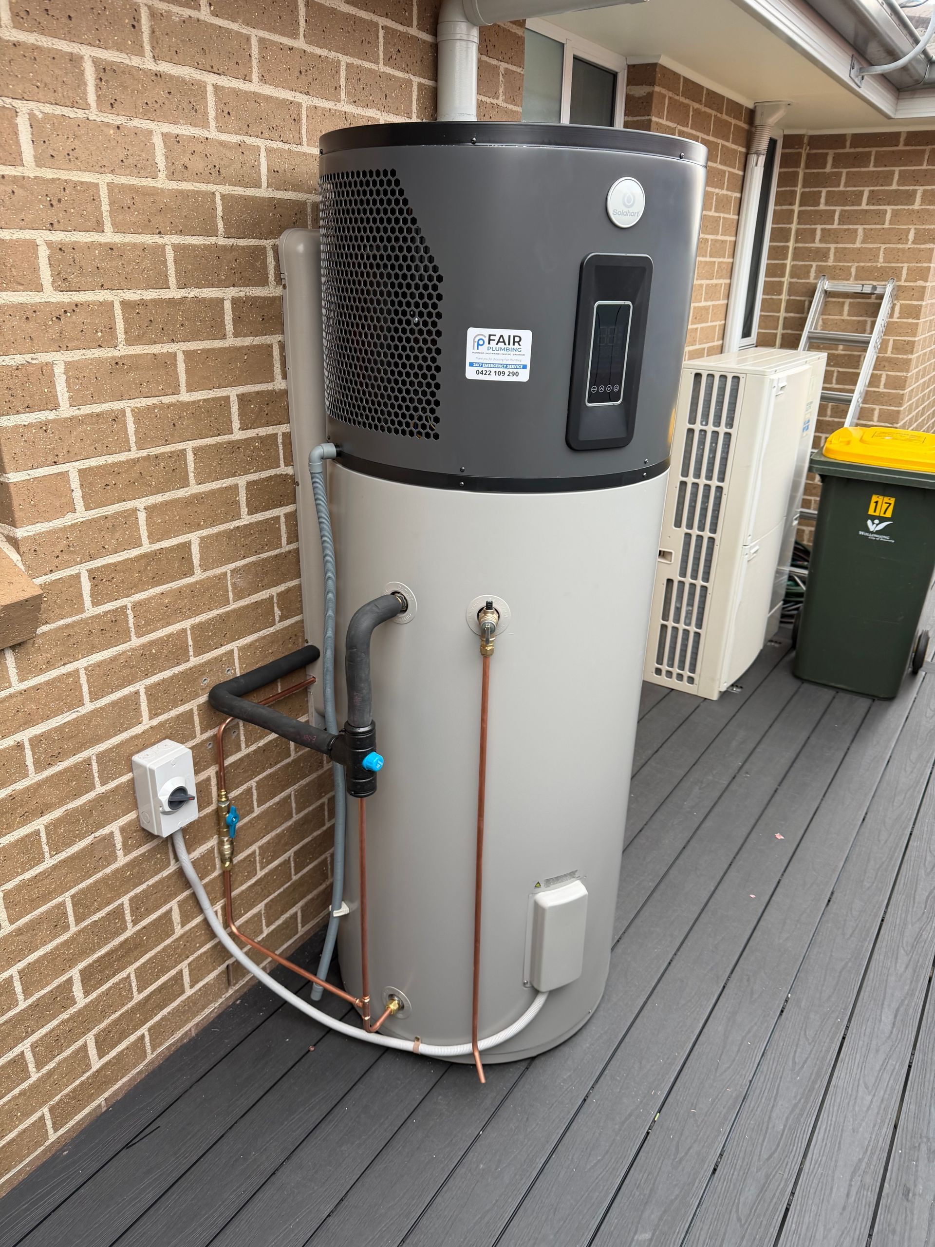 Heat pump water heater on a deck next to a brick wall. A/C unit and trash can visible — Fair Plumbing In Shellharbour, NSW