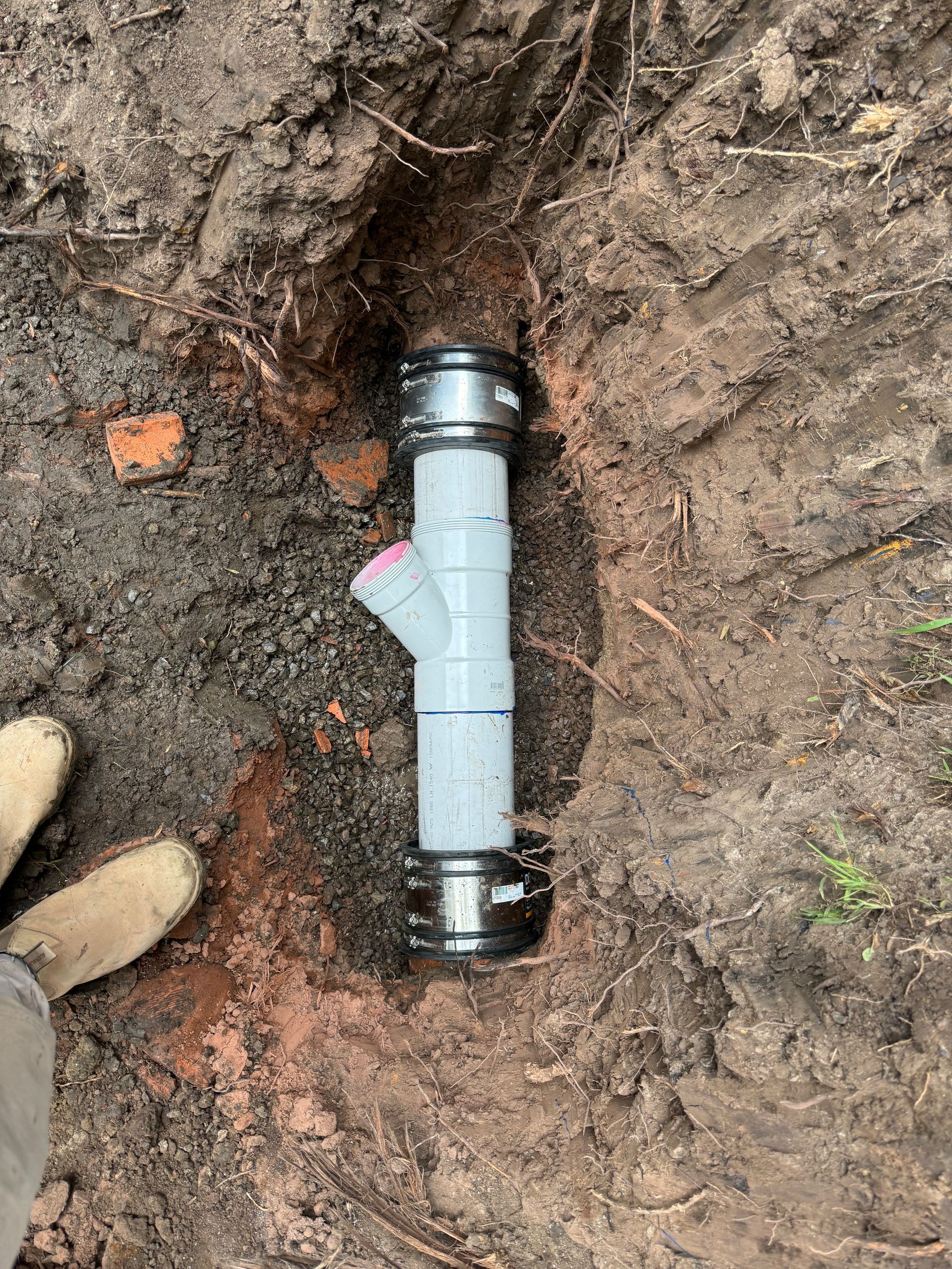 Section of installed plumbing pipe in a dirt trench, featuring gray plastic and metal connections  — Fair Plumbing In Shellharbour, NSW