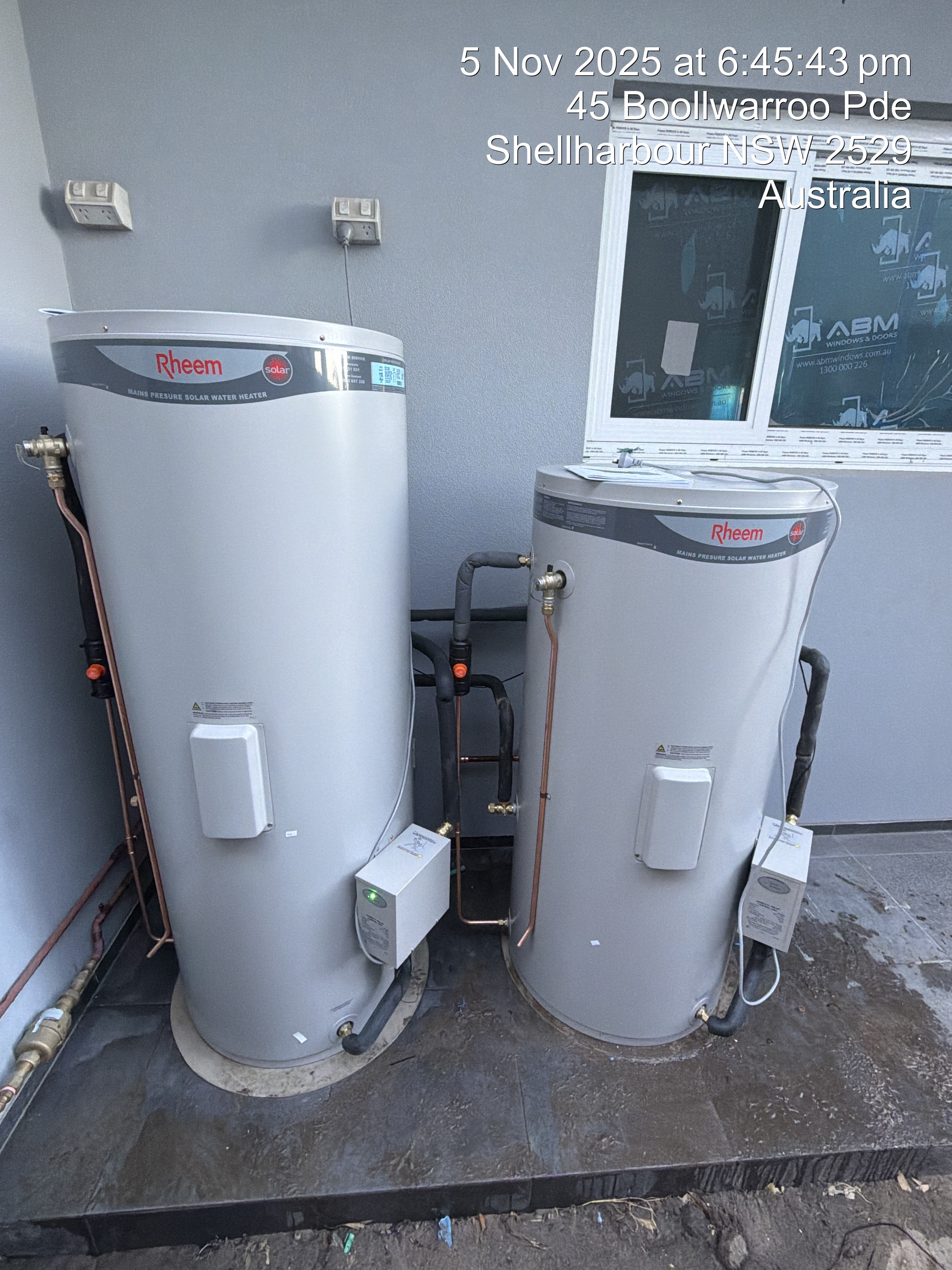 Two silver hot water tanks next to each other on a concrete slab, near a building's wall  — Fair Plumbing In Shellharbour, NSW