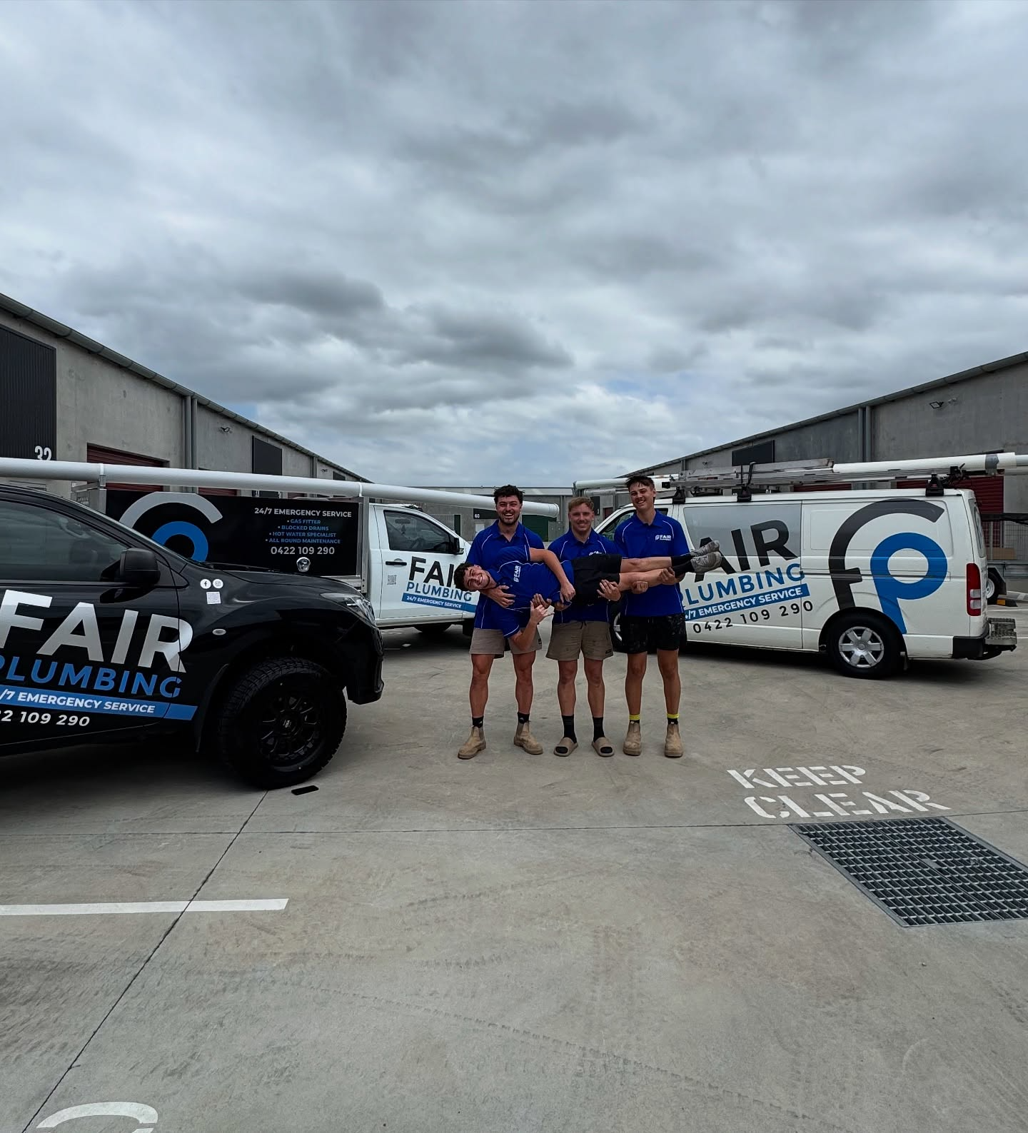 Three plumbers in front of company vans, wearing blue shirts, standing in a parking lot — Fair Plumbing In Shellharbour, NSW