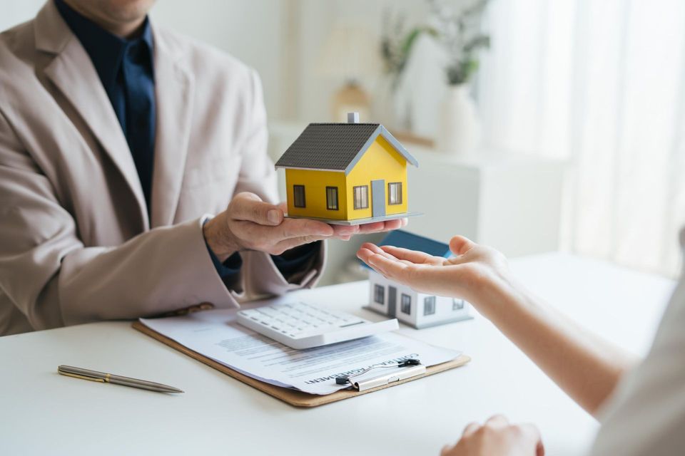 Real estate agent handing a model house to a person at a desk with paperwork and calculator.