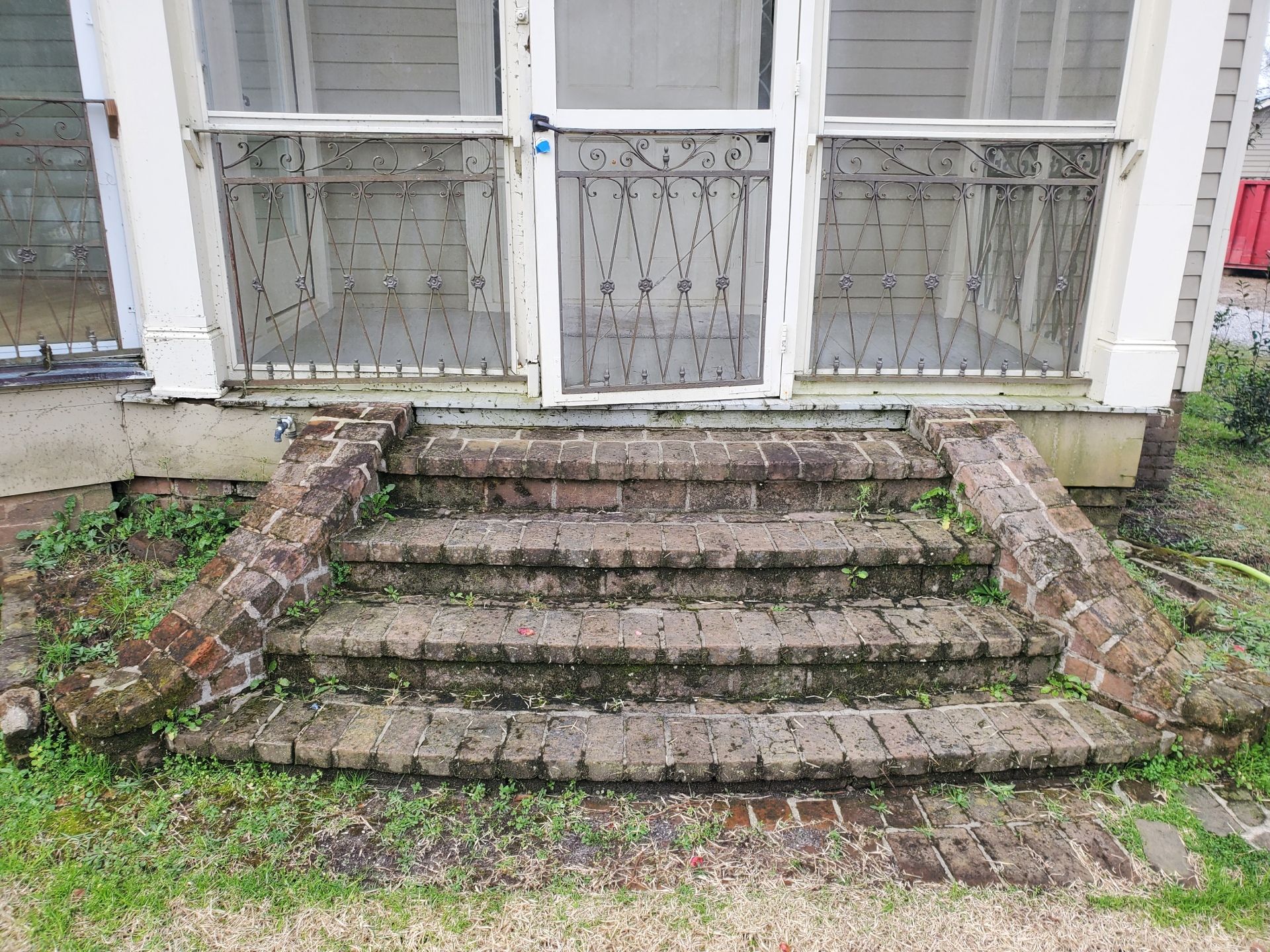 Old brick steps leading up to a screened porch with metal railings, showing signs of weathering and plant growth.