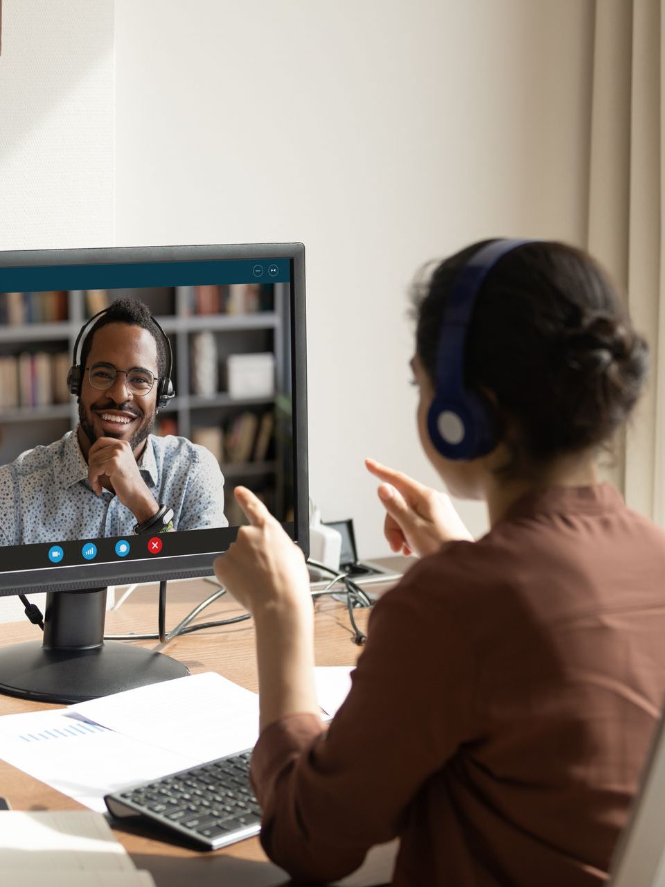 A woman wearing headphones is having a video call with a man on a computer screen.