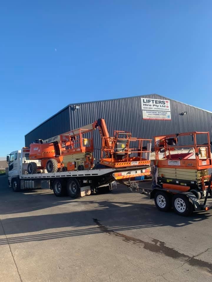Orange Construction Lifts on Flatbed and Trailer Outside a Warehouse Under a Clear Blue Sky — Lifters Hire in Cambridge, TAS