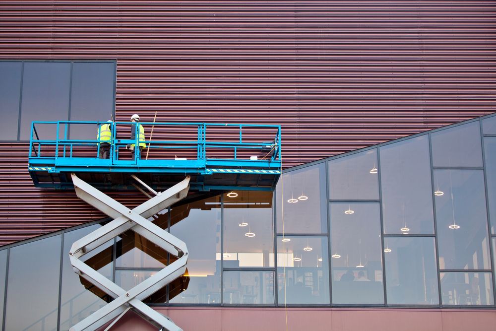 Two Workers on a Blue Scissor Lift Beside a Modern Building With Glass and Brown Siding — Lifters Hire in Cambridge, TAS