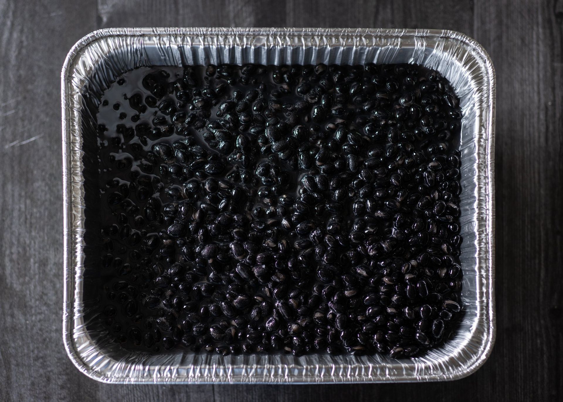 A tin foil pan filled with black beans on a wooden table.