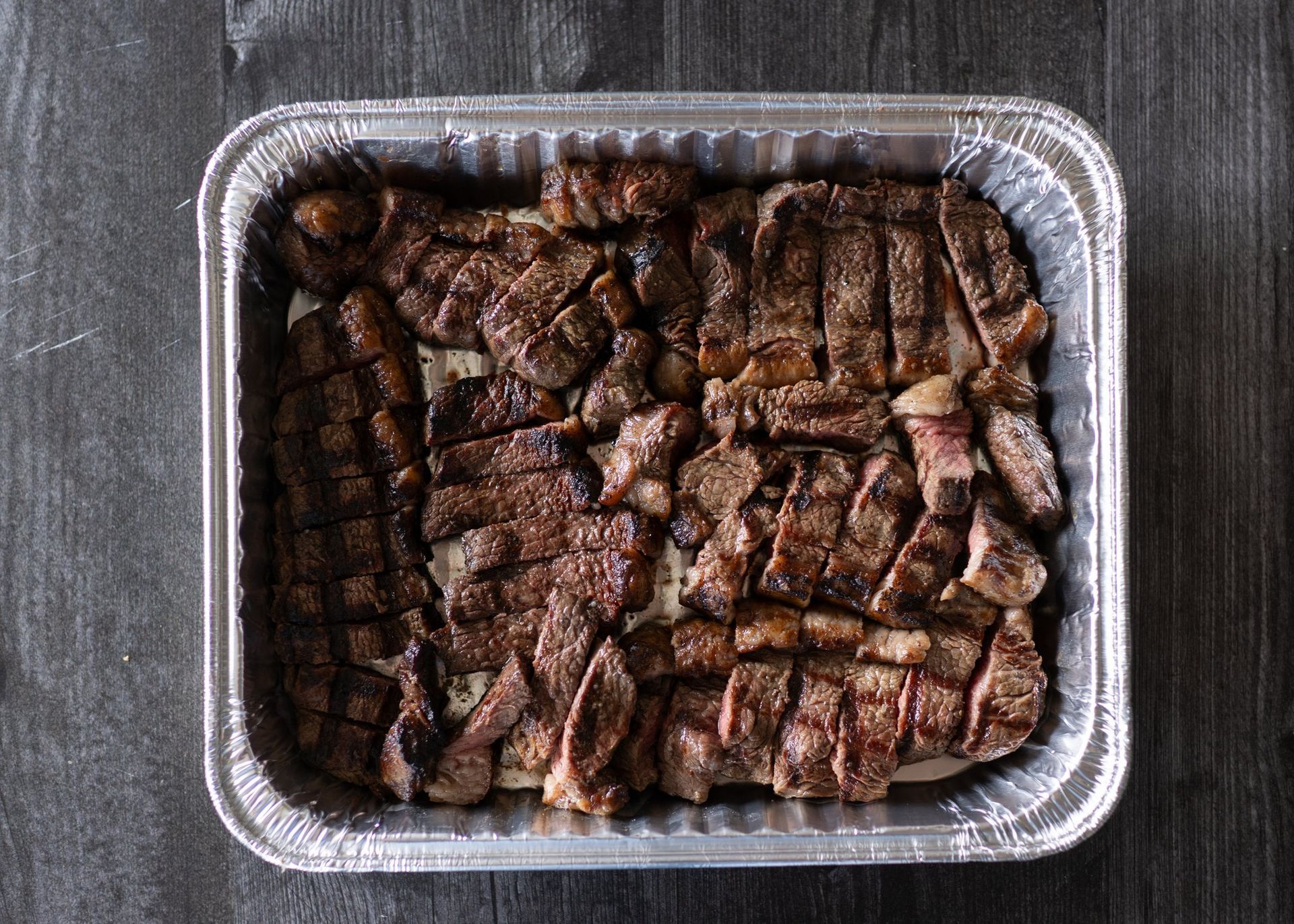 Aluminum foil tray filled with sliced steak on a wooden table.