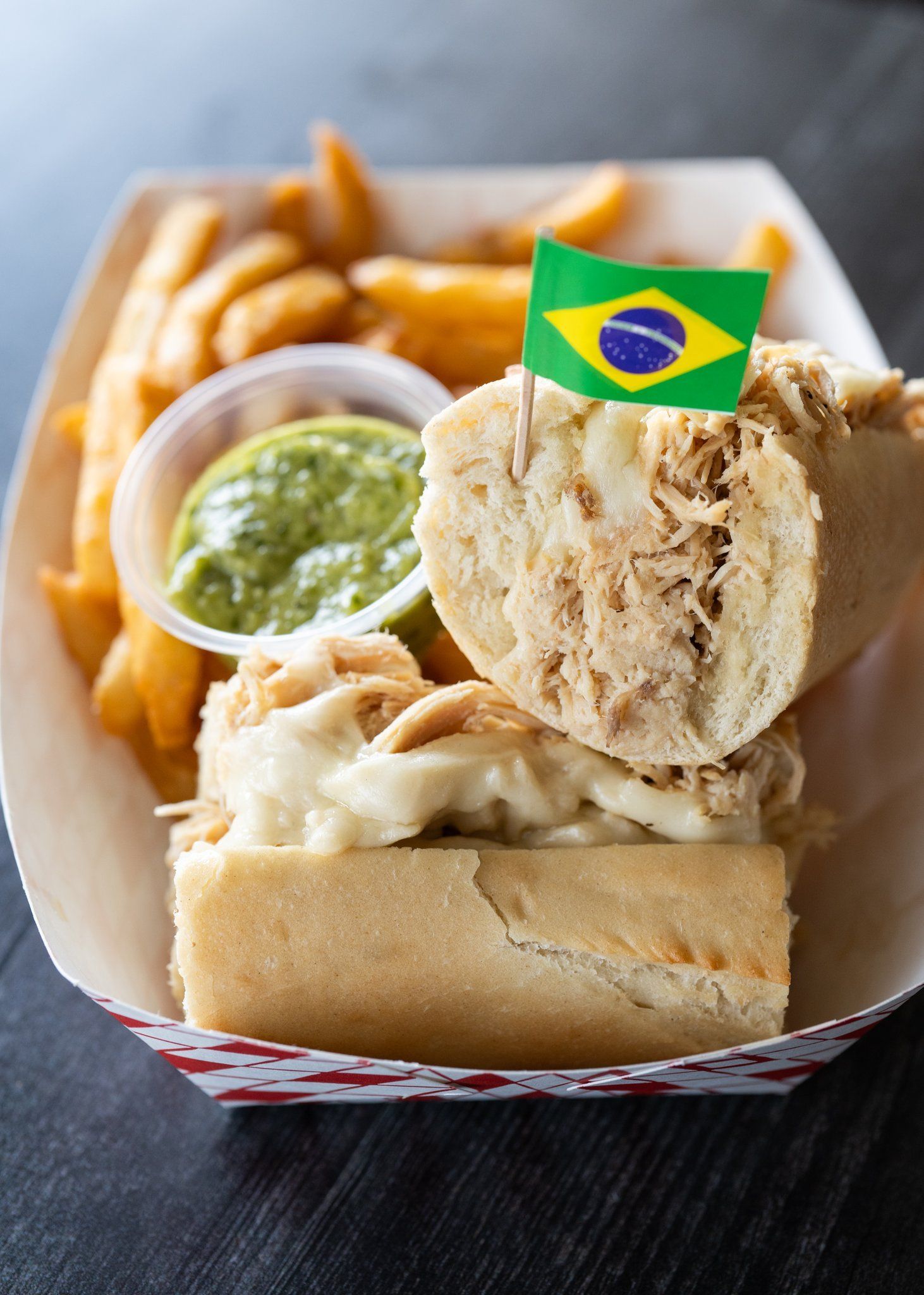 A sandwich with a flag sticking out of it and french fries in a paper container.