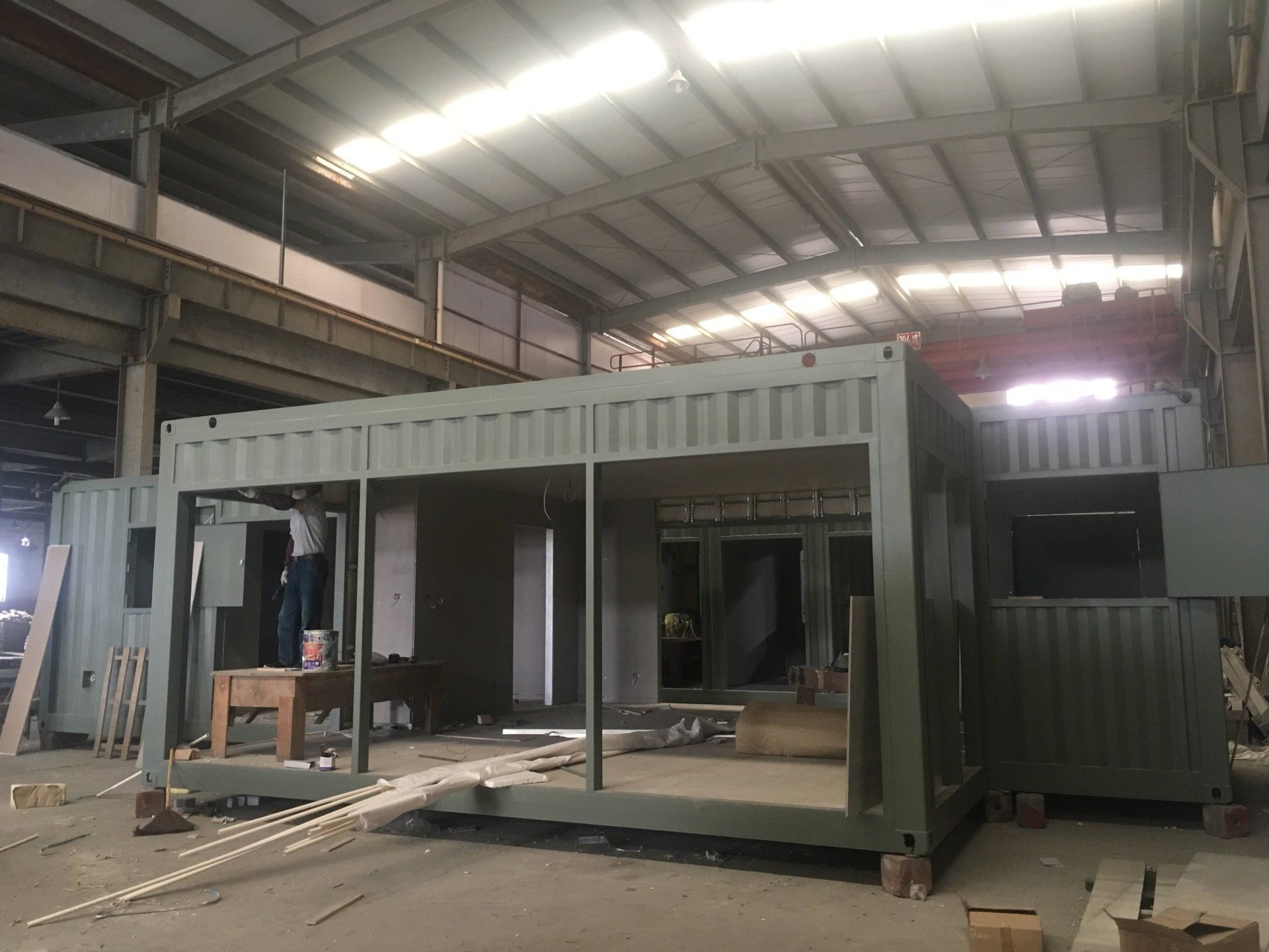 Metal shipping containers being modified in a factory. Structures are olive green with open frames.
