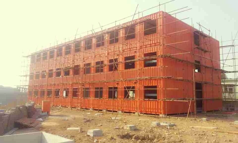 Three-story building under construction; orange shipping containers with window frames, scaffolding surrounding.