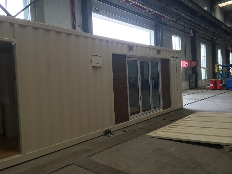 Cream-colored container building with sliding glass doors and brown wooden accents, inside a large warehouse.