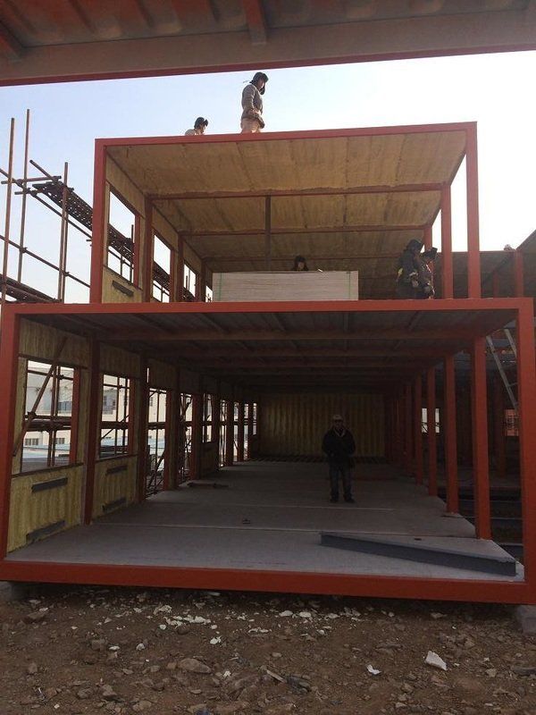 Two-story building under construction; workers on site. Red metal frame with open windows and doors.