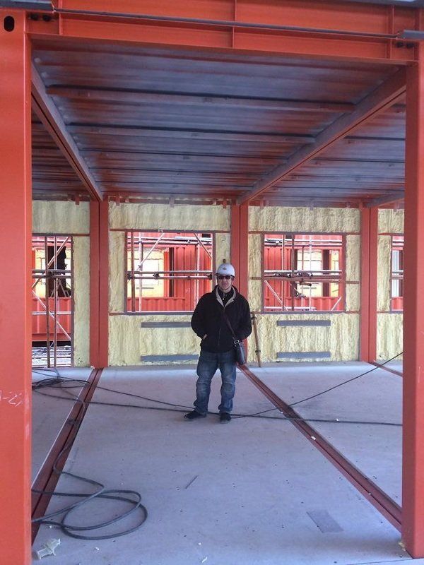 Man in a construction site, standing inside a steel frame. Red beams and metal roof.