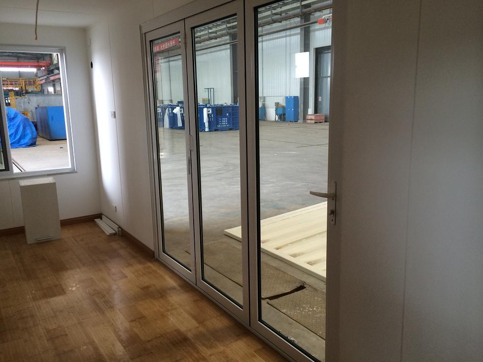 Interior with wood floor, white walls, and large glass folding doors reflecting a factory interior.