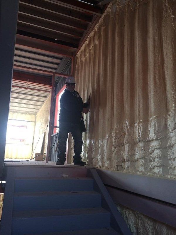 Person insulating a wall inside a building. Beige foam covers most of the wall; a wooden frame is above.