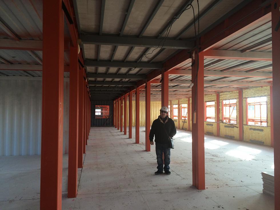 Construction site interior: man standing in a hallway framed by orange support beams and a gray ceiling.