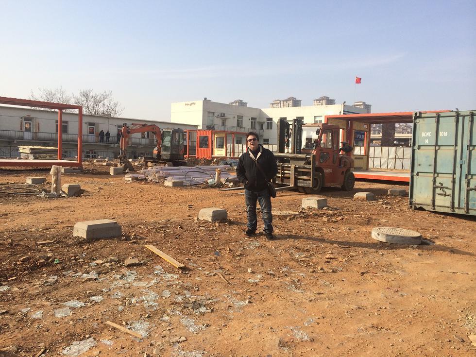 Man standing in a construction site with heavy machinery, orange and blue containers. Sunny day.