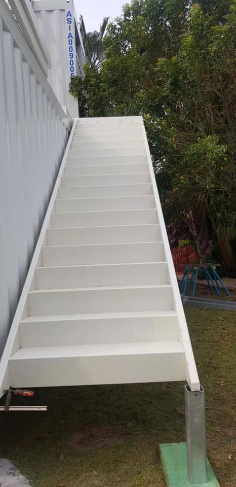White staircase leading up, attached to a white building. Green grass and foliage surround the stairs.