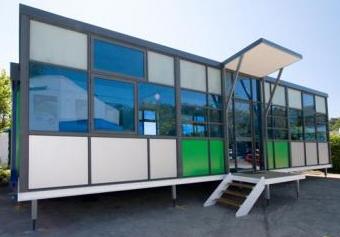 Modern, colorful, glass-walled building with a white porch and steps, raised on metal legs.