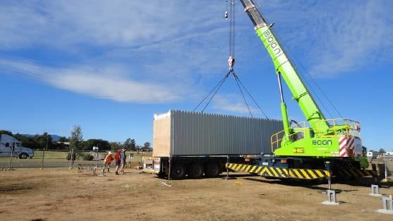 Green crane lifting a rectangular structure from a semi-trailer on a construction site; two people watch.