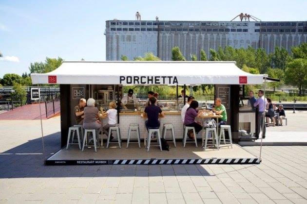 Outdoor restaurant Porchetta, with customers at bar. White awning, stools, and building in background.