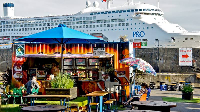 Food stall made from shipping container, blue umbrella, outdoor seating, cruise ship in background.