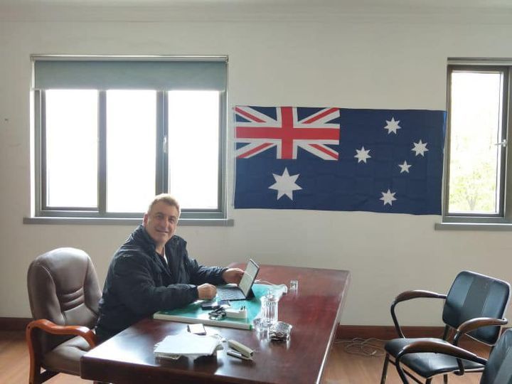 Man sits at desk with laptop in front of an Australian flag.