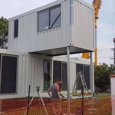 Two-story container home under construction; white containers, windows, workers, tools, and a crane.