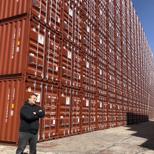 Man standing in front of stacked, reddish-brown shipping containers on a sunny day.