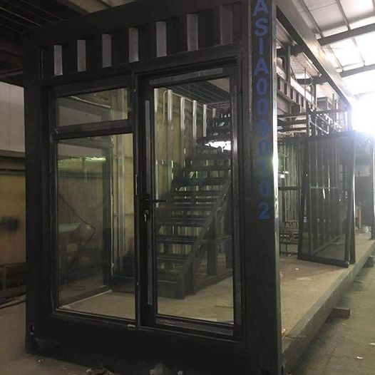Metal structure with glass windows and door, interior staircase under construction.