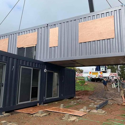 Construction site: crane lifting a shipping container with plywood-covered window cutouts onto another container.
