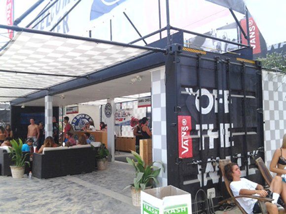Vans retail space built from a shipping container. People relax outdoors. Black structure with checkered awning.
