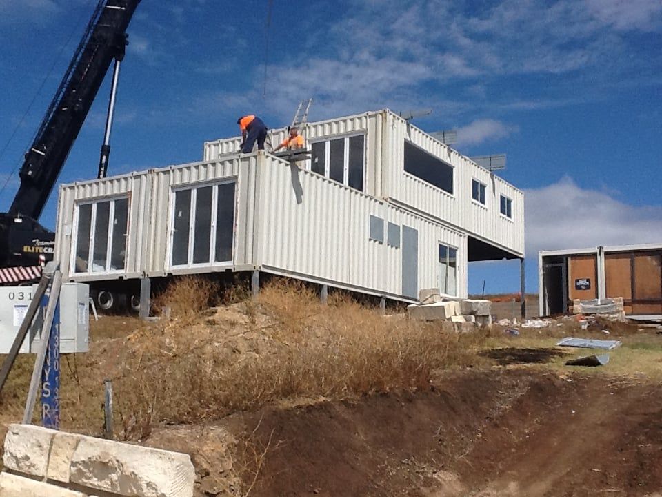 Construction workers placing a shipping container on top of a house; crane in use; blue sky.