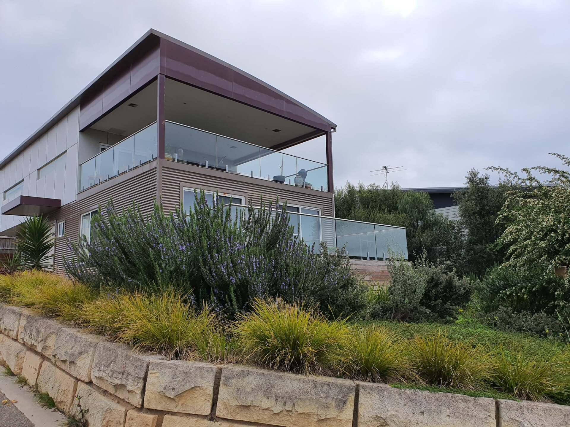 Modern two-story building with glass balconies, brown roof, and lush landscaping on a hillside under a cloudy sky.