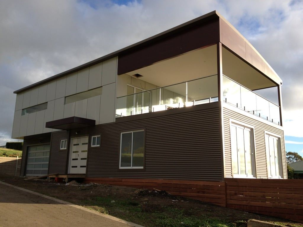 Modern two-story house with brown and white corrugated metal siding, large windows, and a balcony.