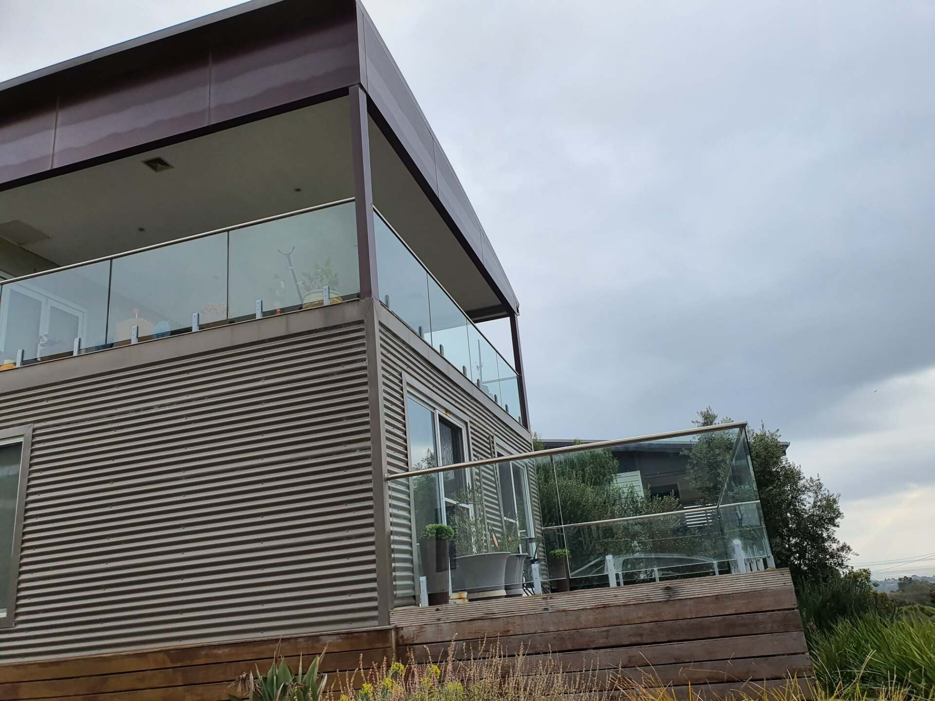 Modern house with glass railings on a deck. Corrugated metal siding, wood deck, and cloudy sky in background.