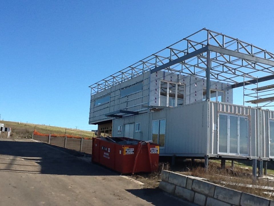 Building under construction, with metal frame on top of repurposed shipping containers, red dumpster on side.