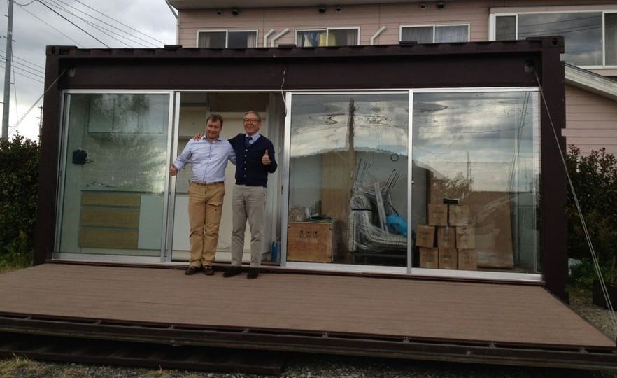 Two men stand in front of a container home with glass doors. One gives a thumbs up. A deck is in front.