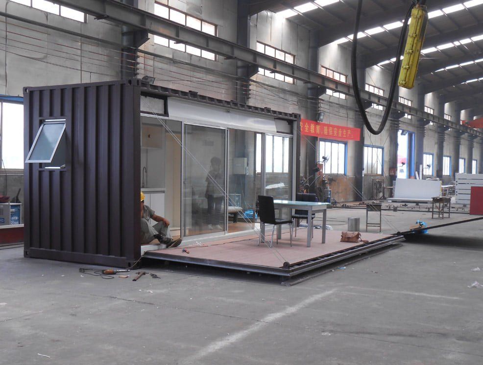Brown shipping container modified into a building with glass walls, outdoor deck, and a person working inside a factory.