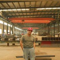 Man in a hard hat poses in a large industrial workshop. Overhead crane visible.