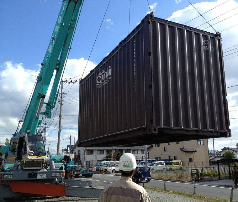 Crane lifting a dark brown shipping container outdoors on a sunny day. A person in a hard hat watches.