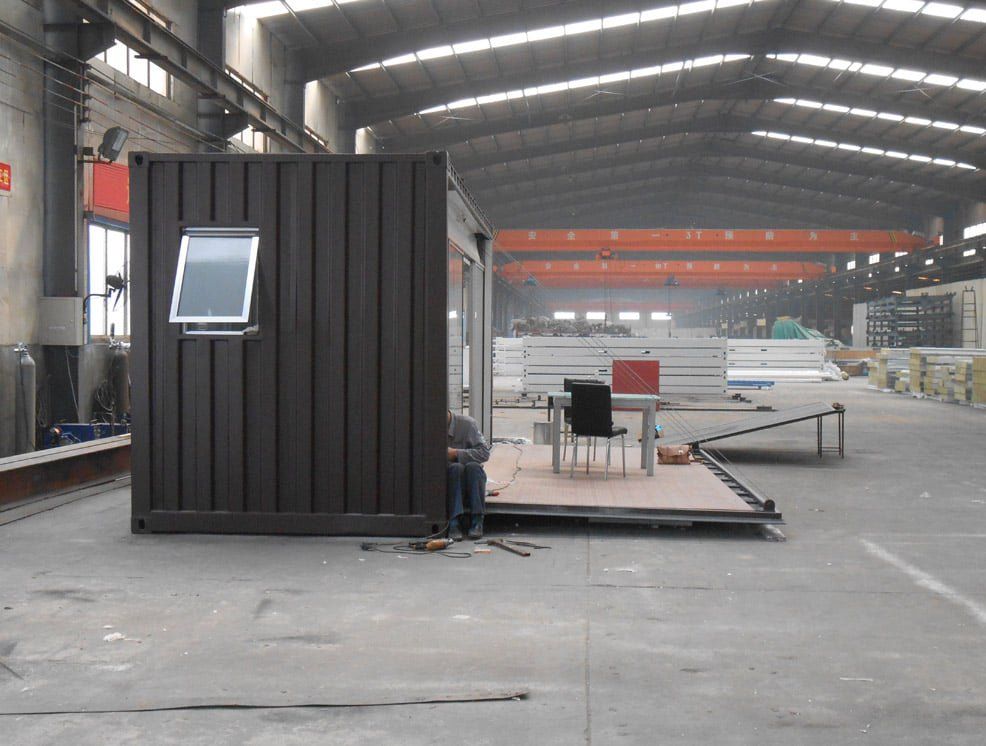 Dark brown shipping container with window, open door with table and chair, person working. Factory setting.