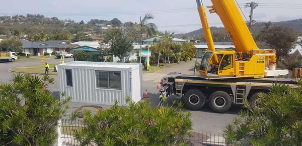 A yellow crane lifting a white container with windows in a residential area.