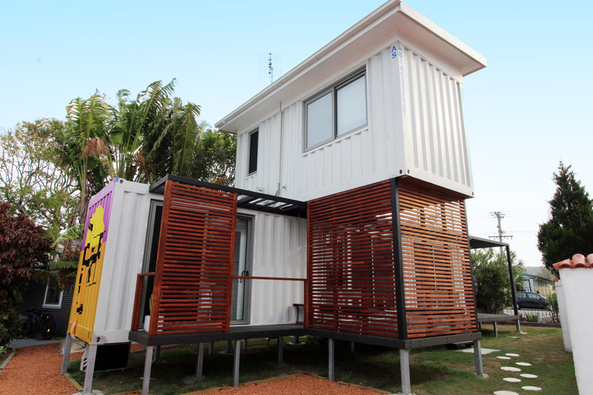 Two-story white container home with wooden deck and slatted facade, set in a yard with trees.