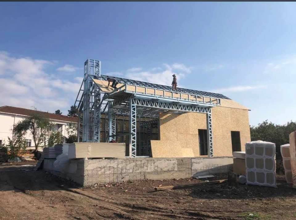 Construction of a building with a metal frame. Workers on the roof, OSB walls, and foundation visible. Sunny day.