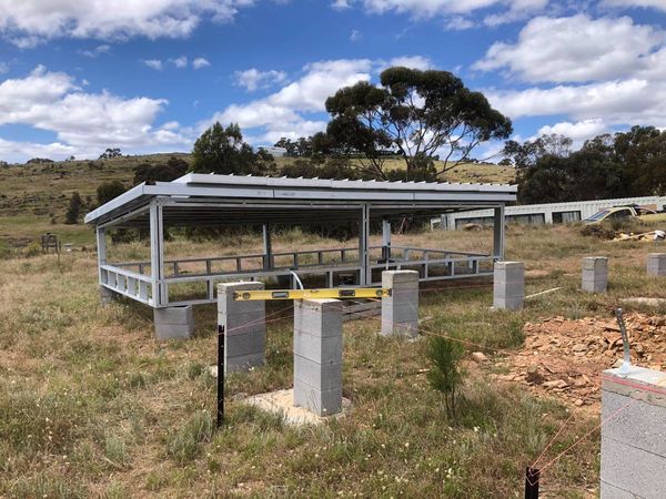 Metal structure under construction with a roof, sitting on concrete blocks, in a grassy field.