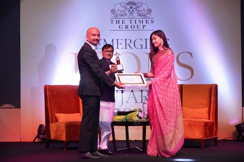 Man in suit receives award from a woman in a pink saree on stage at an event.