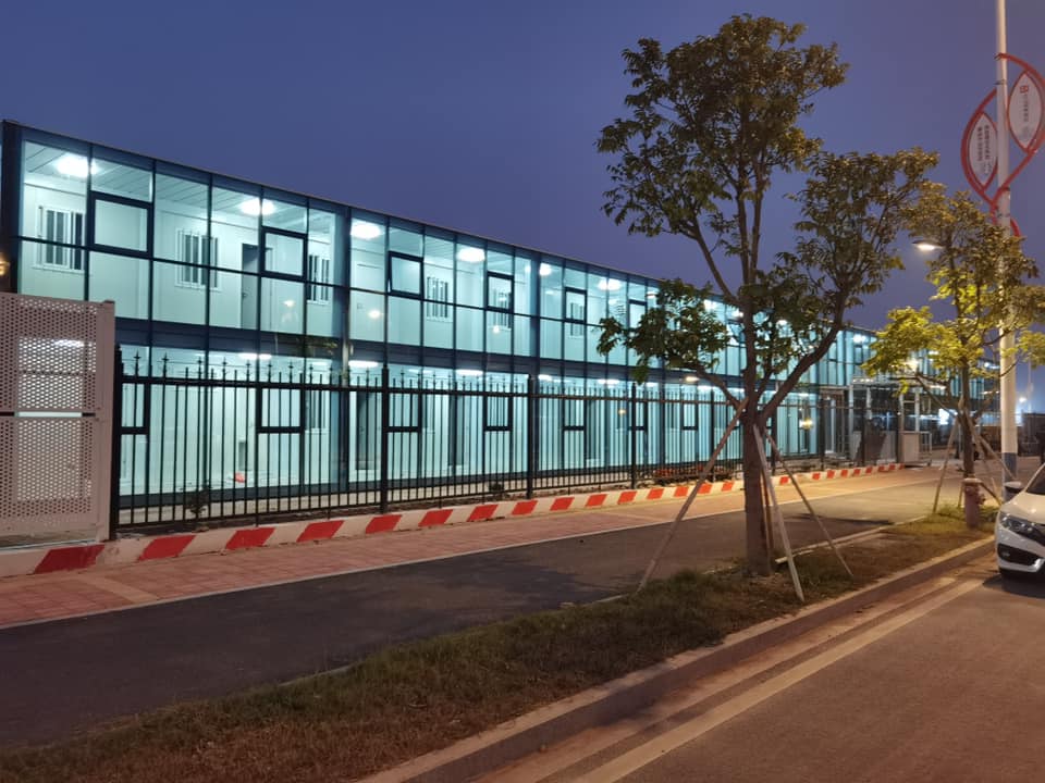 Two-story modular building with glass walls, fenced perimeter, and street, at dusk.