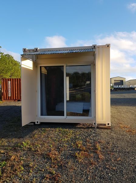White shipping container converted into a small structure, with a sliding glass door and an awning, outdoors.