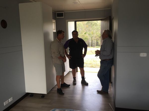 Three men standing inside a gray room with an open doorway to the outside.